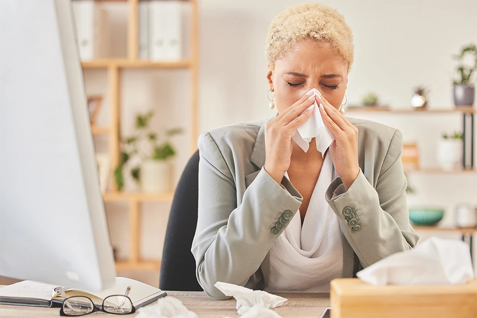 Woman experiencing Allegra side effects, possibly allergic rhinitis, blowing her nose in an office setting.