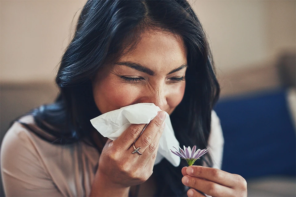 Fexofenadine provides relief for a woman with allergies as she sneezes into a tissue while holding a small flower.
